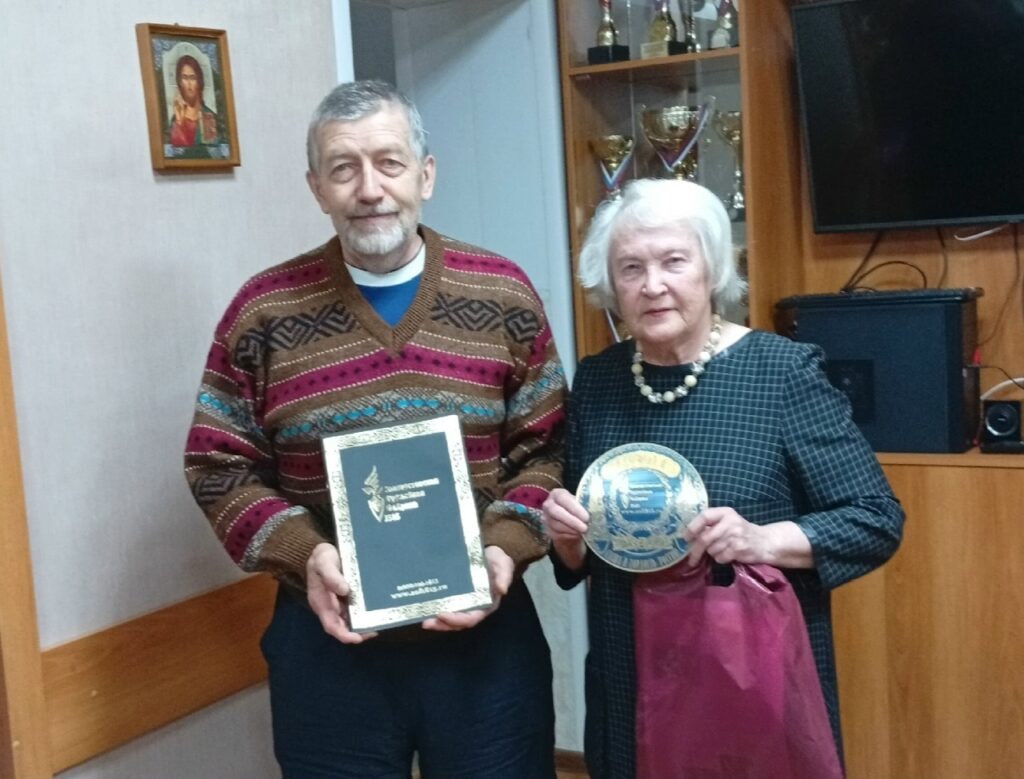 Пожилые мужчина и женщина стоят в комнате с кубками; он держит папку/книгу, она — памятную тарелку и бордовый пакет, рядом на стене телевизор и икона.