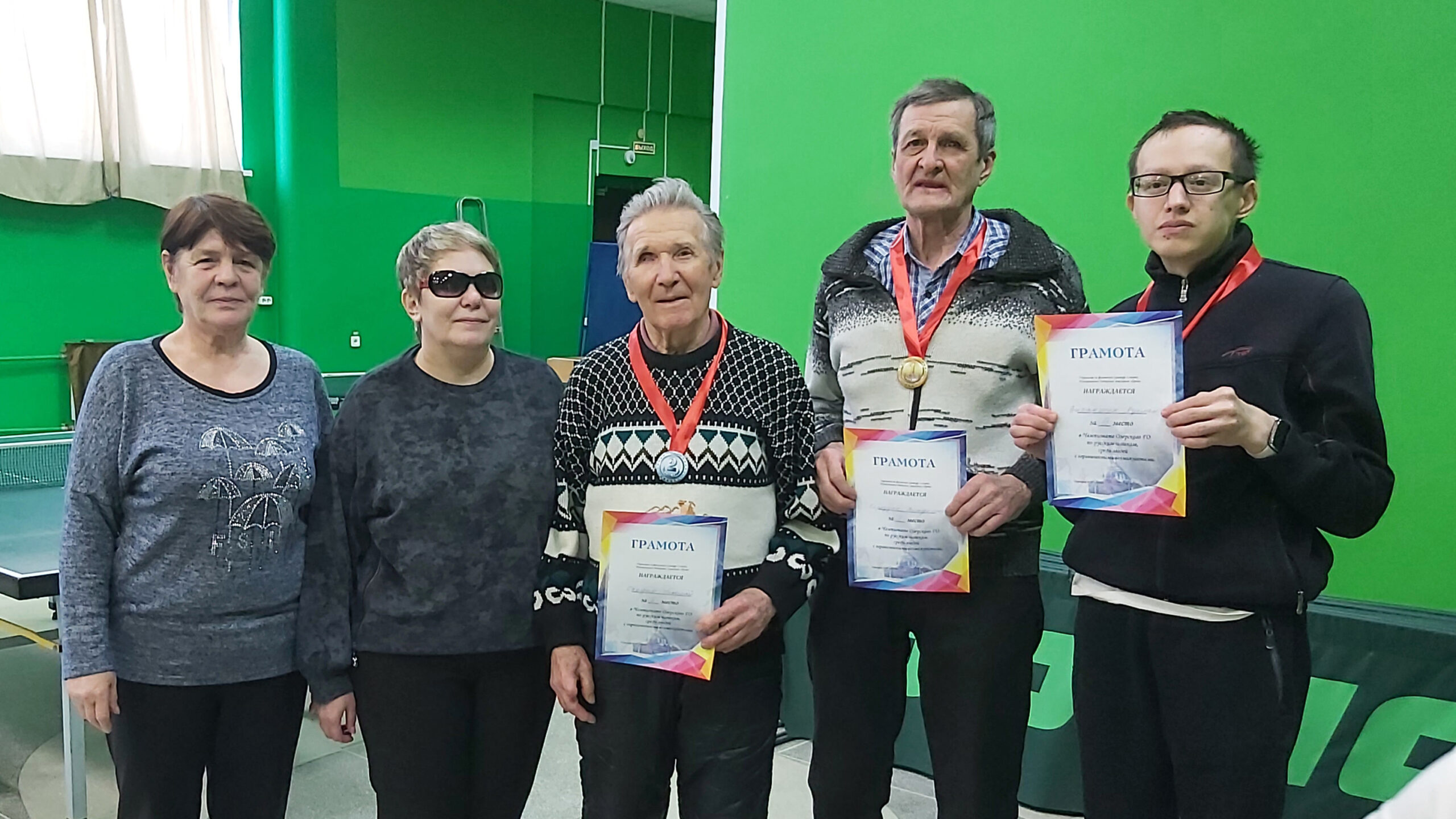 Пятеро людей в зале для настольного тенниса; трое держат дипломы «ГРАМОТА», у двоих видны медали на красных лентах. Слева водяной знак «Samsung Triple Camera / Снято на Galaxy A70 Наталья М».