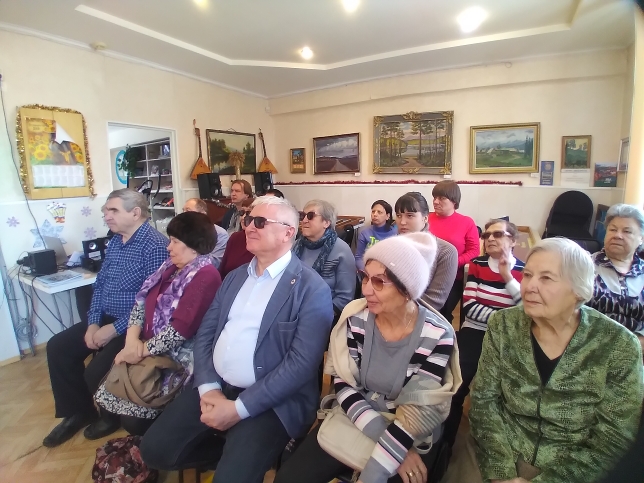 Пожилые люди сидят рядами в комнате с картинами на стенах и слушают рассказ библиотекаря