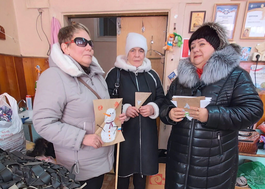 Три женщины в зимних пуховиках стоят в помещении и держат в руках картонные поделки: слева аппликация со снеговиком, у двух других треугольные заготовки. На стене сзади висят рамки с грамотами/дипломами.