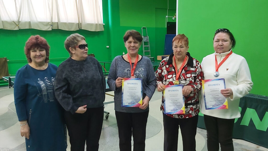 Пять женщин в зале с зелёными стенами; трое держат грамоты «ГРАМОТА» и медали на красных лентах. На заднем плане теннисный стол и окна с шторами.