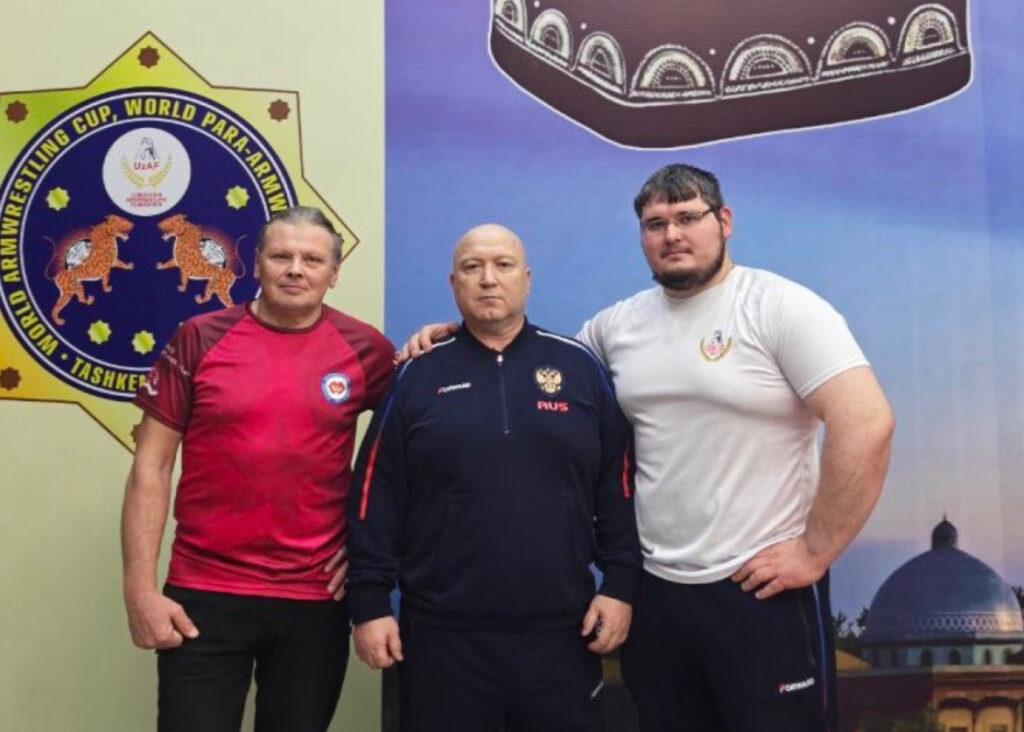 Трое мужчин на фоне баннера соревнований по пара-армрестлингу: слева в красной форме, в центре в темном костюме с надписью RUS, справа в белой футболке