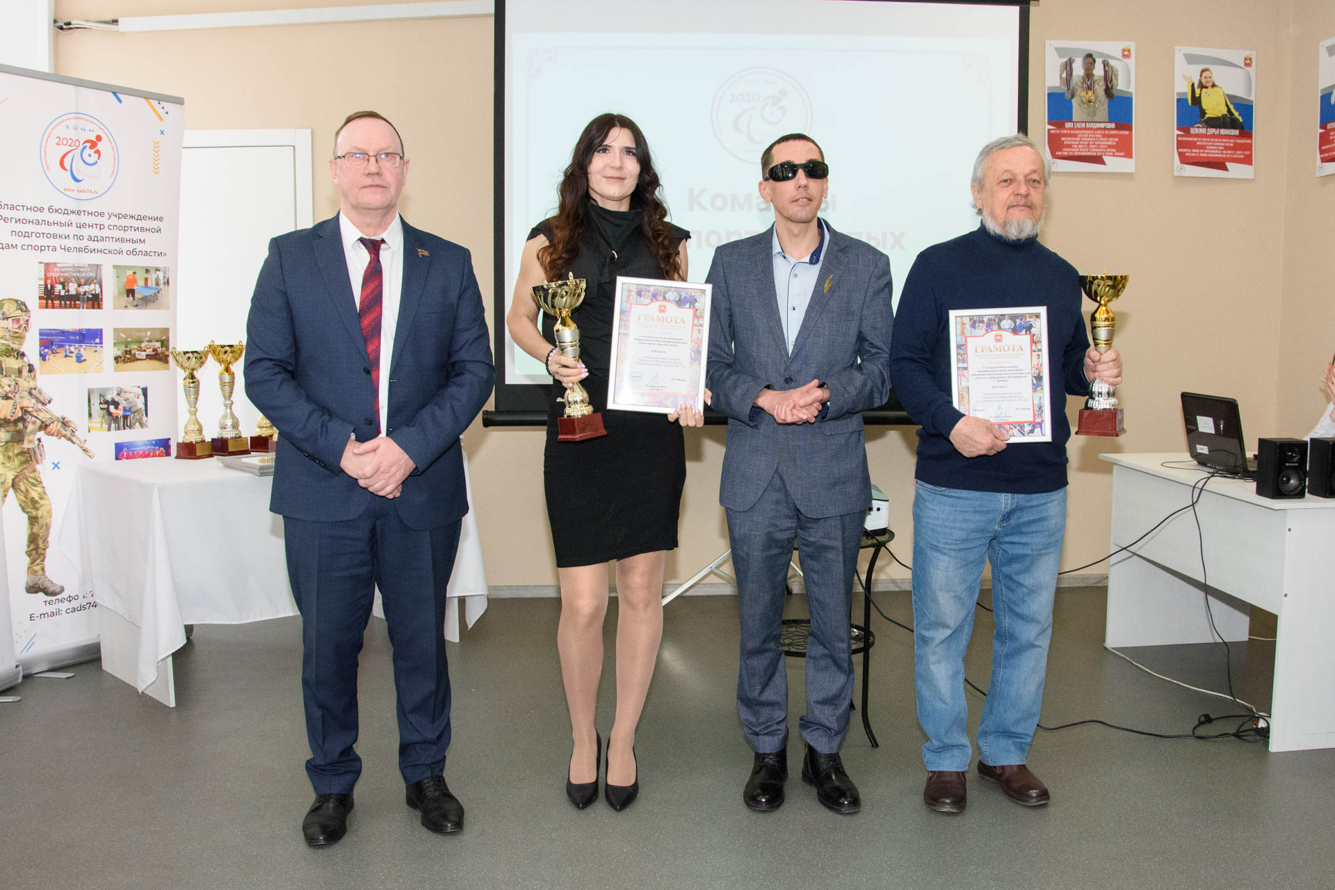 Четыре человека на церемонии награждения в помещении: двое держат кубки и дипломы «ГРАМОТА», на столе рядом ещё несколько кубков, на фоне экран проектора.