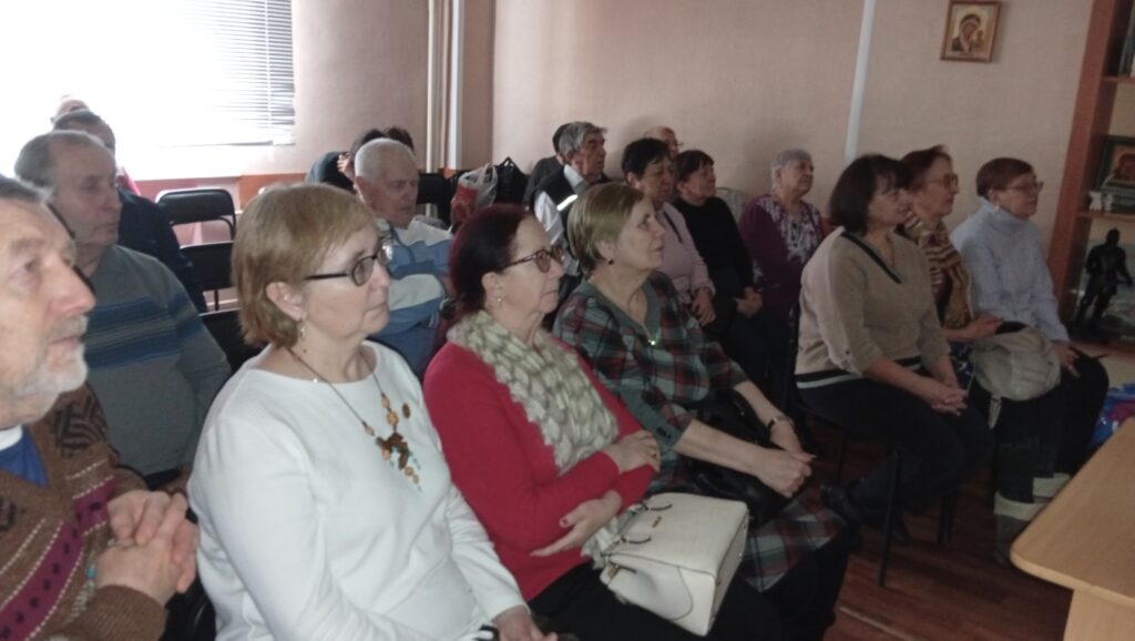 Пожилые люди сидят рядами в комнате и внимательно слушают в одну сторону. Слева окно с жалюзи, справа полки и икона на стене.