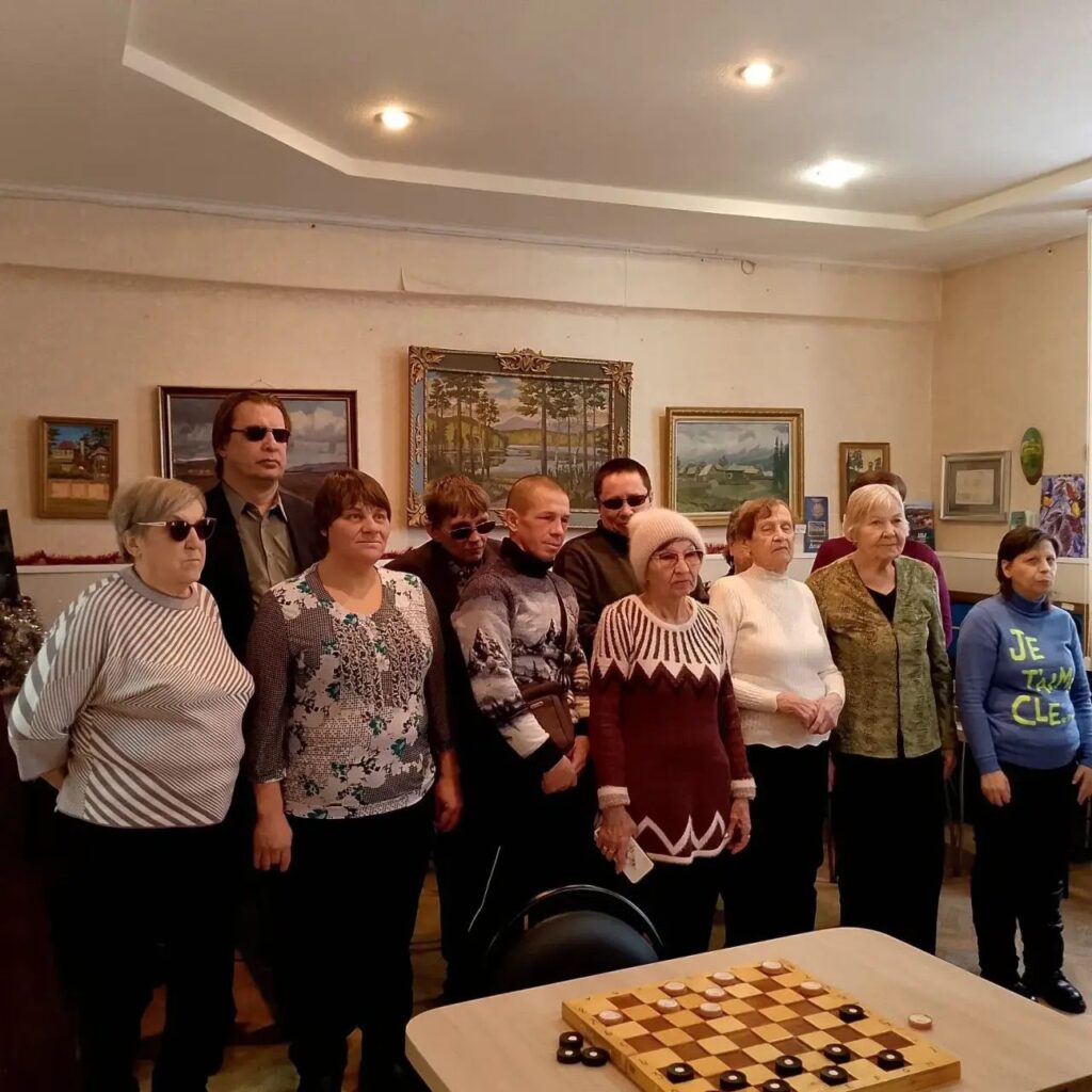 Группа людей стоит в комнате перед стеной с картинами. На переднем плане стол с шашечной доской.