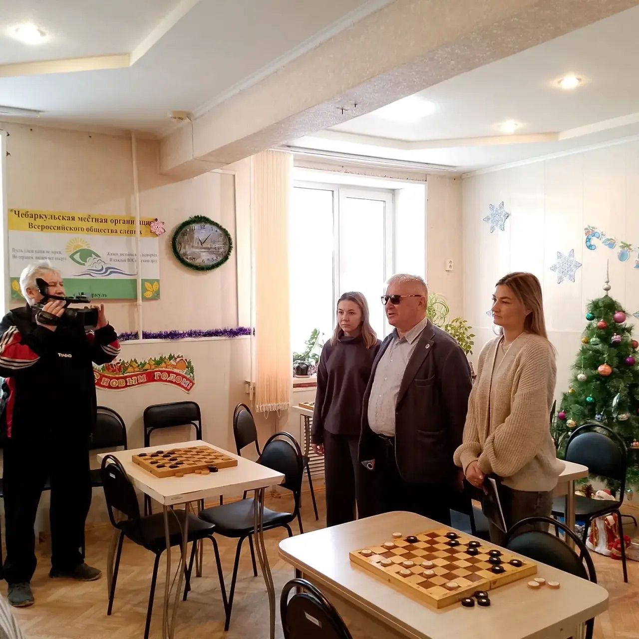 Комната с шашечными досками на столах. Слева мужчина снимает на камеру. У окна стоят трое людей. На стенах новогодние украшения и плакат общества слепых, справа украшенная ёлка.