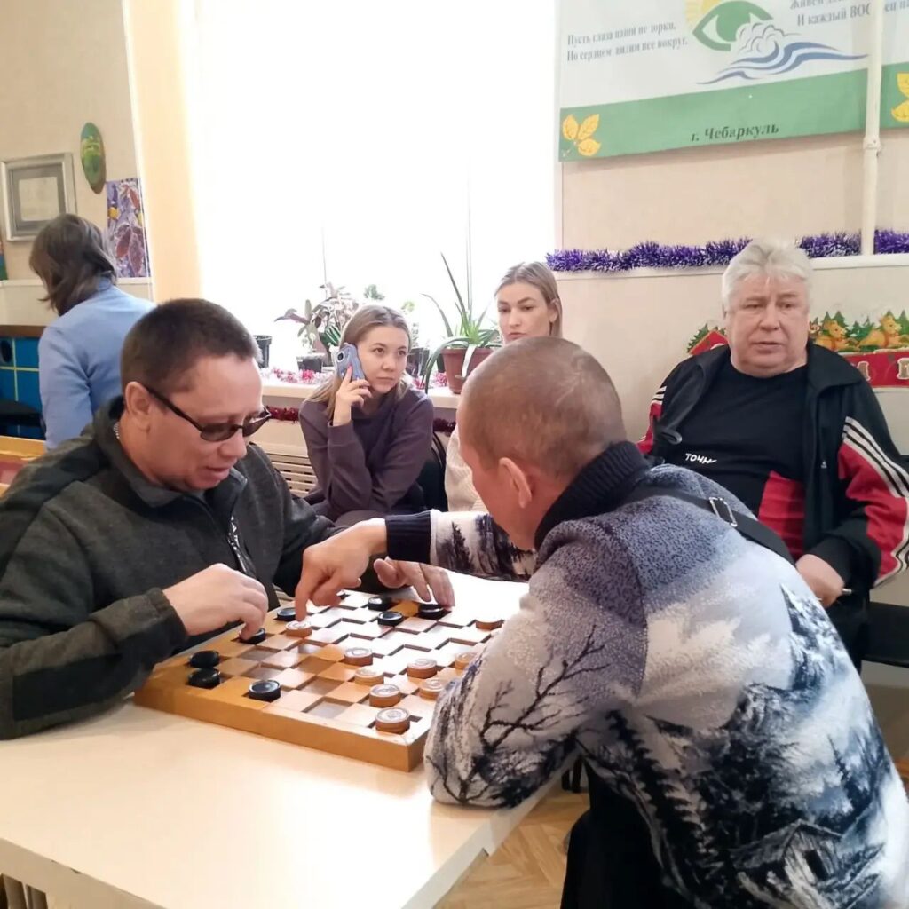 Двое мужчин за столом играют в шашки, один делает ход. На заднем плане сидят люди, две девушки и пожилой мужчина; одна девушка держит телефон у уха.