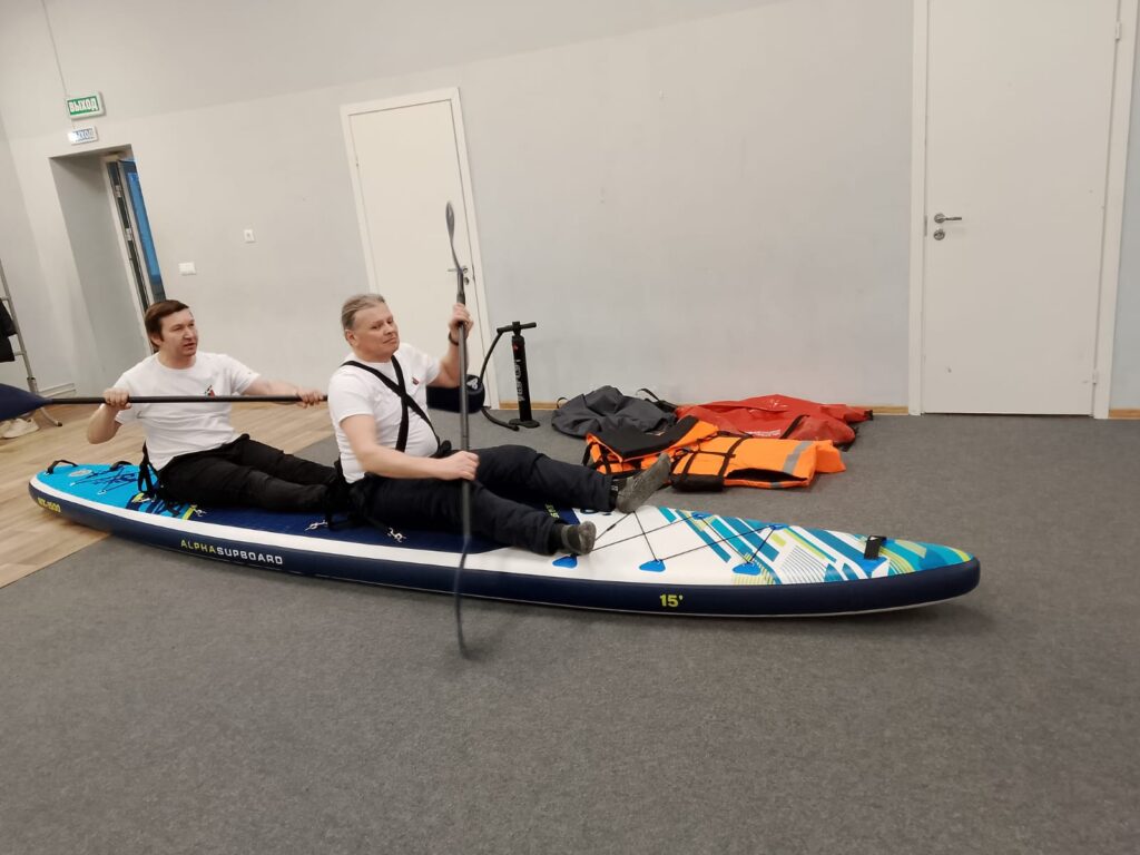 двое мужчин сидят на надувной SUP-доске и держат вёсла. Рядом на полу оранжевые спасжилеты и насос.