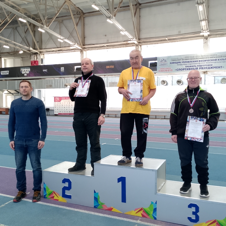 Внутри спортивного зала: трое мужчин с медалями и грамотами стоят на пьедестале 1–3 (в центре на месте 1), рядом слева стоит ещё один мужчина без медали.