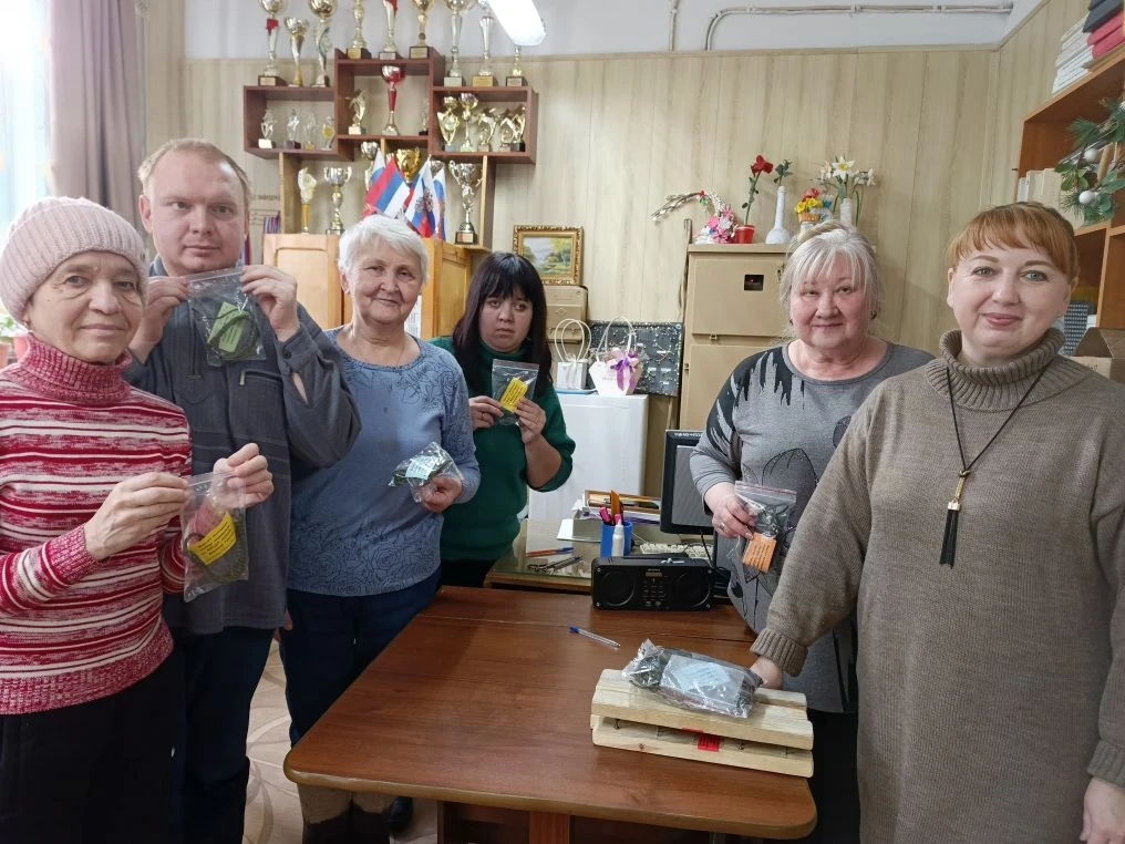 Шесть участниц мастер-класса в комнате стоят у стола и держат небольшие прозрачные пакетики с предметами. На заднем плане полки с кубками и флажками.