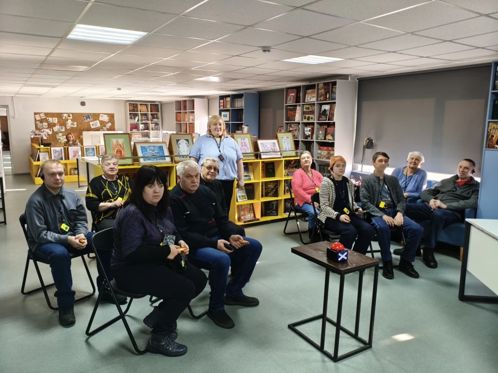 команды сидят и слушают вопросы. позади стеллажи с книгами и рамками с рисунками. Впереди маленький столик с двумя круглыми предметами (красный с белым крестом и синий с белыми линиями).