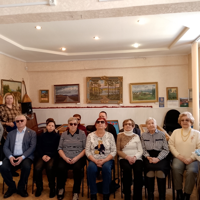 ряд из семи сидящих пожилых людей; за ними на стене несколько пейзажей в рамах, сверху потолок с точечными светильниками. Слева у стены стоит женщина; рядом заметны колонка и балалайка.