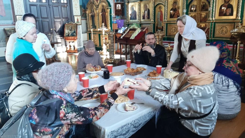 Люди в православном храме сидят за столом с выпечкой и красными стаканчиками; две женщины передают друг другу кусок торта, на фоне иконостас.