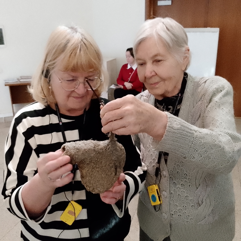 Две пожилые женщины держат и рассматривают большой серо-коричневый шероховатый предмет; у обеих на шее жёлтые бейджи.