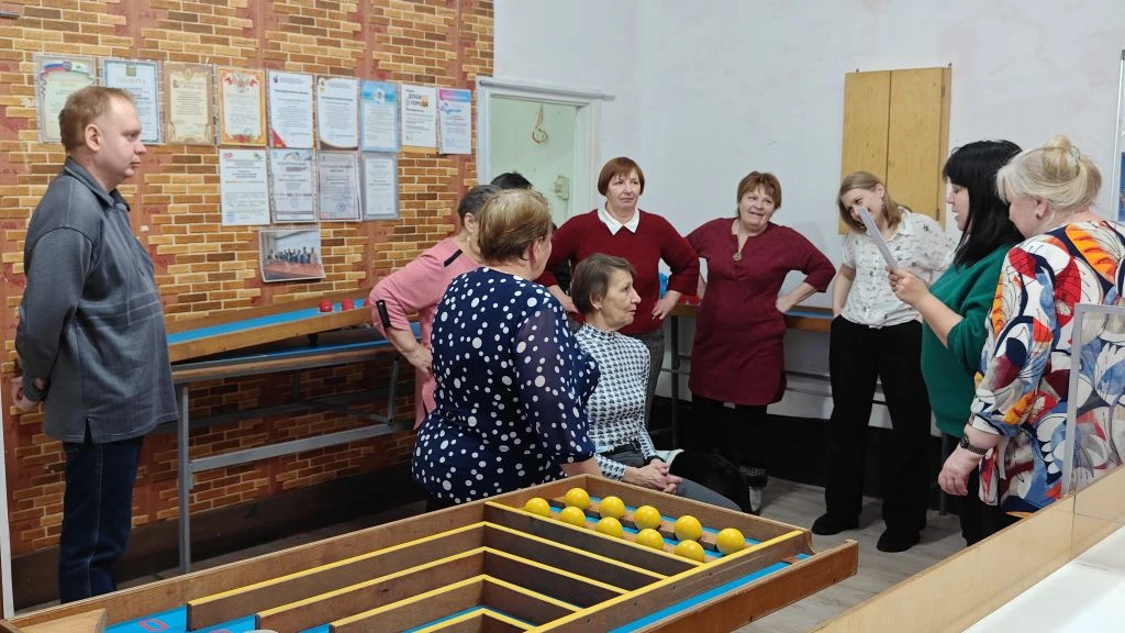 В комнате стоит группа людей, одна женщина читает с листа. На переднем плане деревянный игровой стол с жёлтыми шариками; на стене развешаны документы.