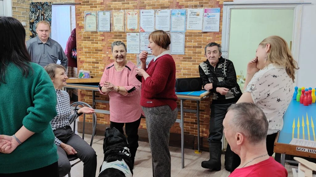 В комнате собралась компания людей, они разговаривают и смеются; на полу лежит чёрная собака, справа игровой стол с маленькими кеглями.