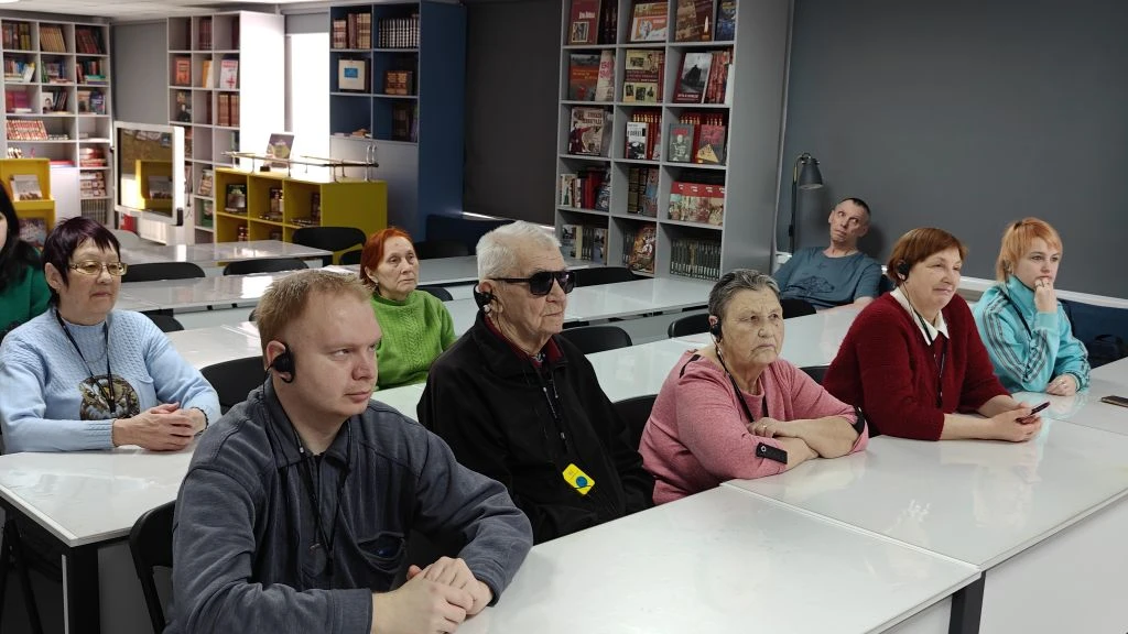 Люди сидят за длинными столами, как на занятии; многие в наушниках. На фоне книжные стеллажи, похоже на библиотеку или учебный класс.