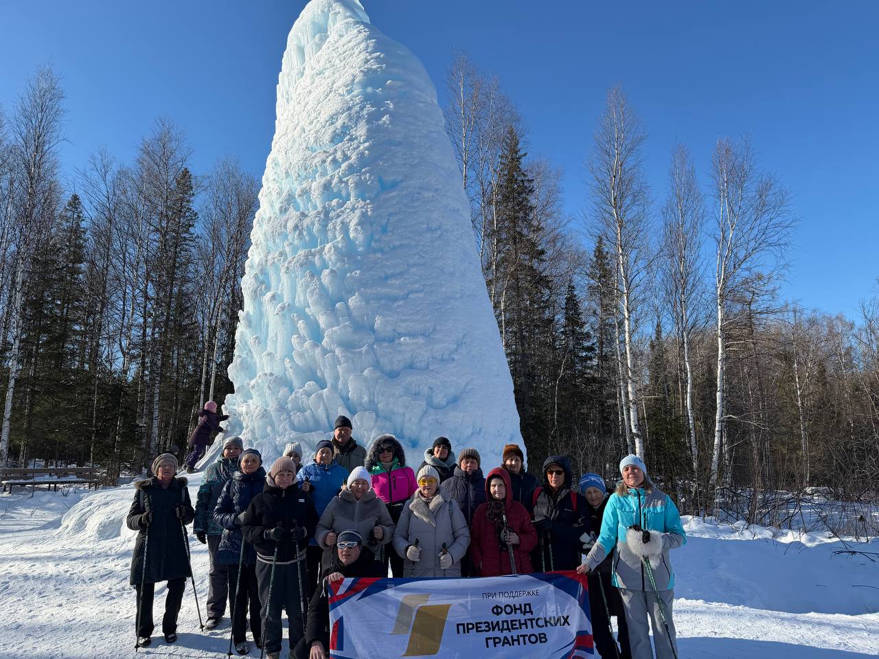 Группа людей в зимней одежде фотографируется на снегу у огромной ледяной глыбы/столба в лесу; спереди держат баннер «Фонд президентских грантов».