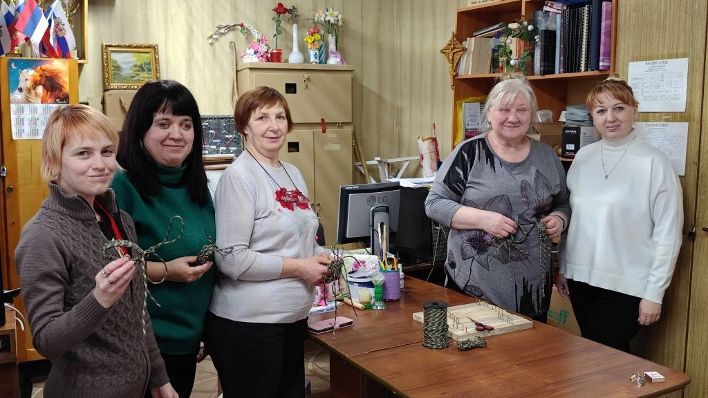 Пять женщин за столом в комнате занимаются рукоделием: плетут/скручивают шнуры на деревянных планках, на столе бечёвка и инструменты.