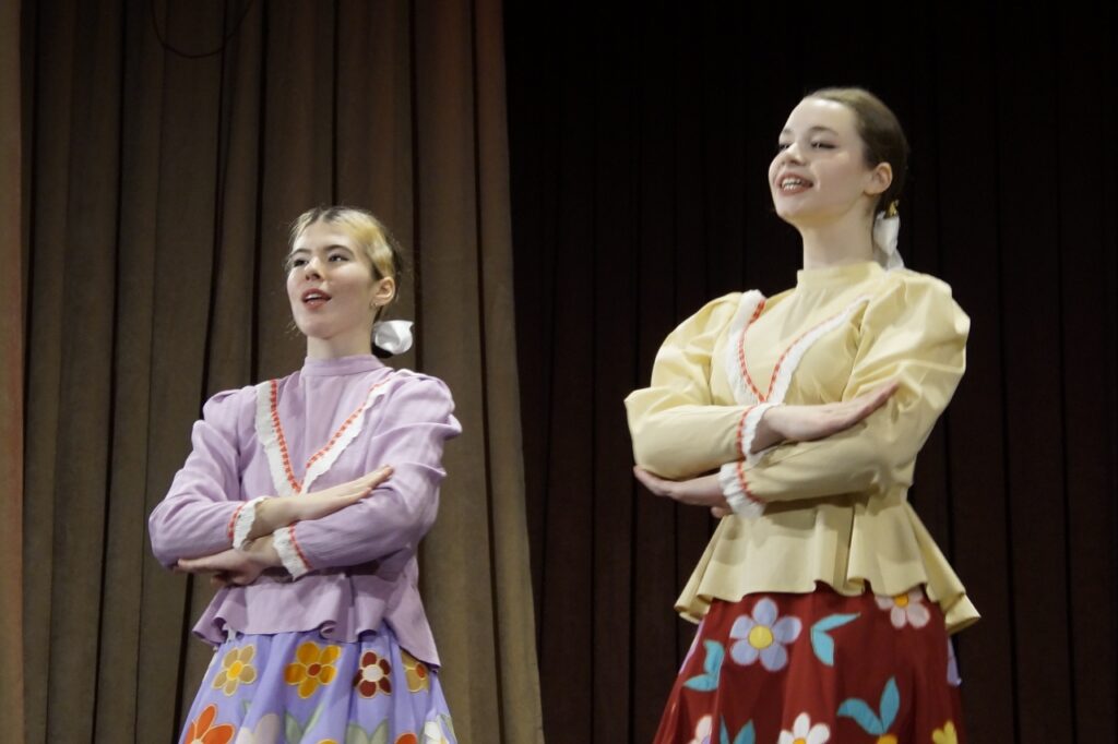 Две девушки на сцене перед тёмным занавесом, стоят со скрещёнными руками и поют/говорят; у обеих банты, одна в сиреневом с цветочной юбкой, другая в светло-жёлтом с красной цветочной юбкой.