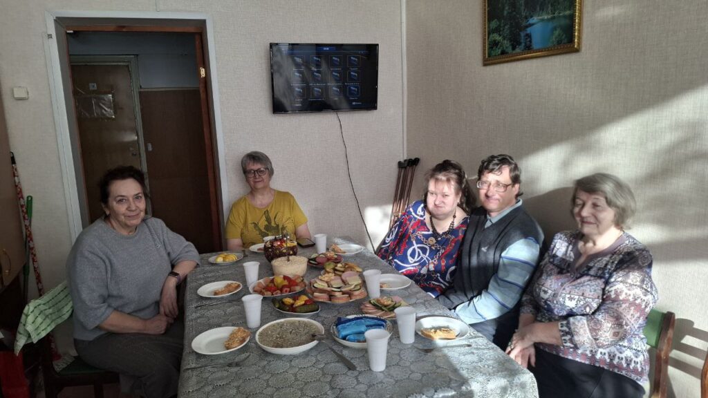 Пять человек сидят за накрытым столом с едой и напитками, позируют для фото; на стене телевизор, справа яркий солнечный свет.