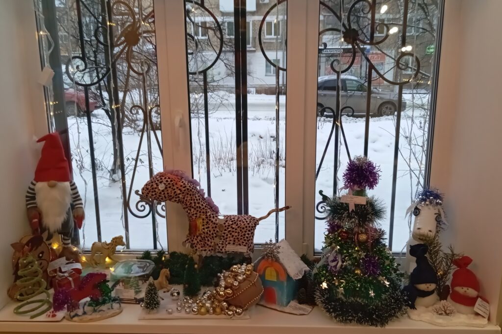 На подоконнике новогодняя композиция: гном, пятнистая лошадка, маленькая ёлка с украшениями, игрушечный домик, шарики/бусы и гирлянда с тёплыми огоньками.