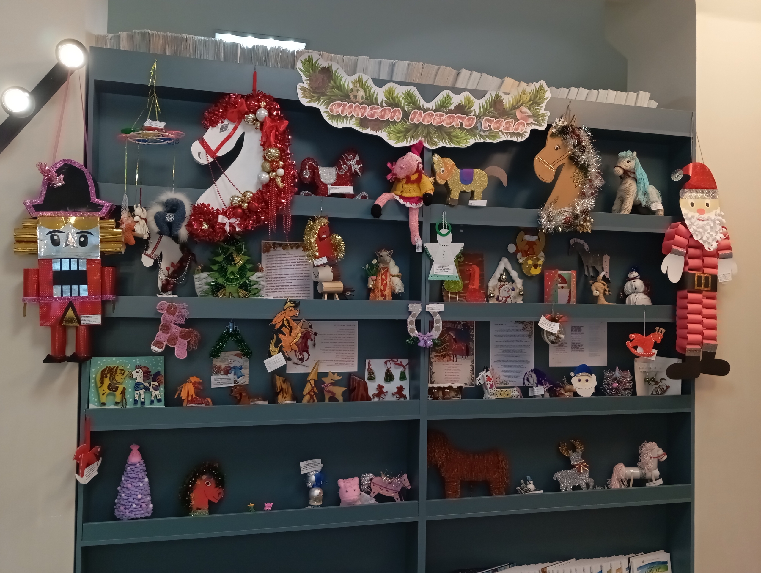 Стеллаж с новогодними поделками: сверху надпись «Счастья нового года!», по полкам много игрушек и украшений, слева большой щелкунчик, справа большой Дед Мороз.