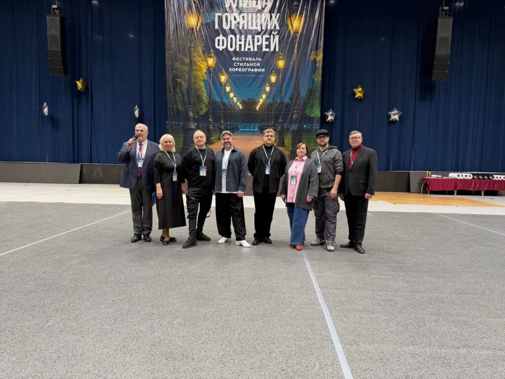 Восемь взрослых стоят на сцене перед баннером «УЛИЦА ГОРЯЩИХ ФОНАРЕЙ» (фестиваль стильной хореографии); слева мужчина с микрофоном, у многих бейджи, по бокам звёздные шары и подвесные колонки.