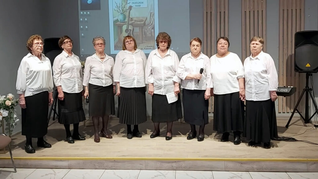 Семь пожилых женщин в одинаковых белых блузках и чёрных юбках стоят на сцене; у одной в руках микрофон, у другой лист бумаги.