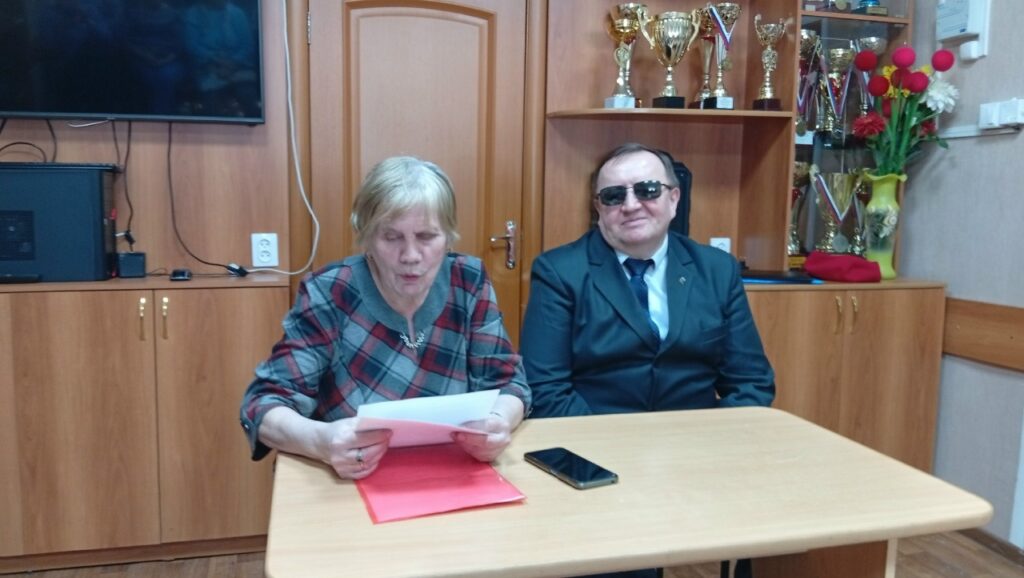 Двое людей сидят за столом: женщина читает лист над красной папкой, рядом лежит смартфон; мужчина в тёмном костюме и тёмных очках. На заднем плане шкаф с кубками и медалями.