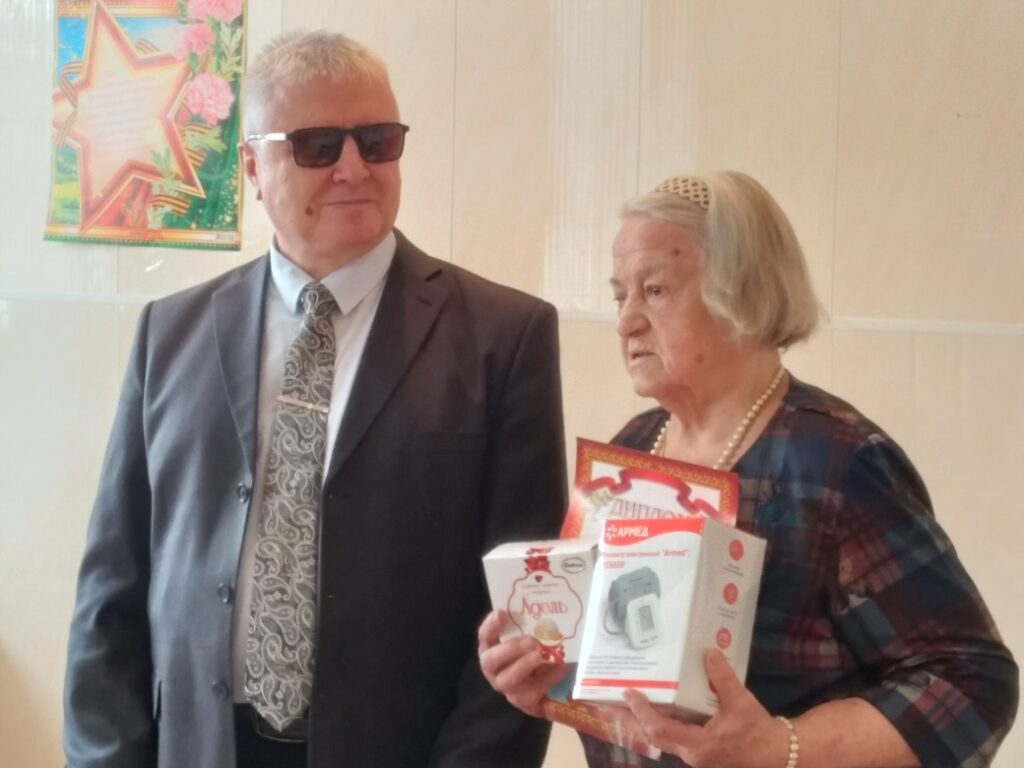 Пожилые мужчина в костюме и тёмных очках и женщина в клетчатом платье с бусами. Женщина держит «Диплом» и коробки «АРМЕД» и «Адель». На стене слева плакат со звездой и цветами.