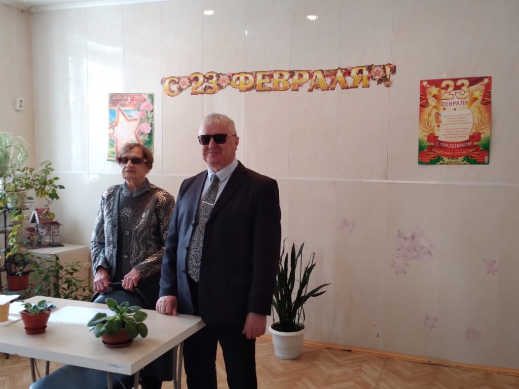 Два пожилых человека в очках стоят у стола в комнате с украшениями к 23 февраля; на стене растяжка «С 23 ФЕВРАЛЯ!» и праздничные плакаты, на столе горшки с растениями и бумаги.