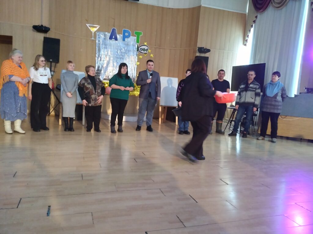 В актовом залеучастники стоят в ряд у сцены; в центре мужчина в костюме с микрофоном, сзади блестящий фон с надписью «АРТ». Спереди проходит женщина с красным тазом/миской.