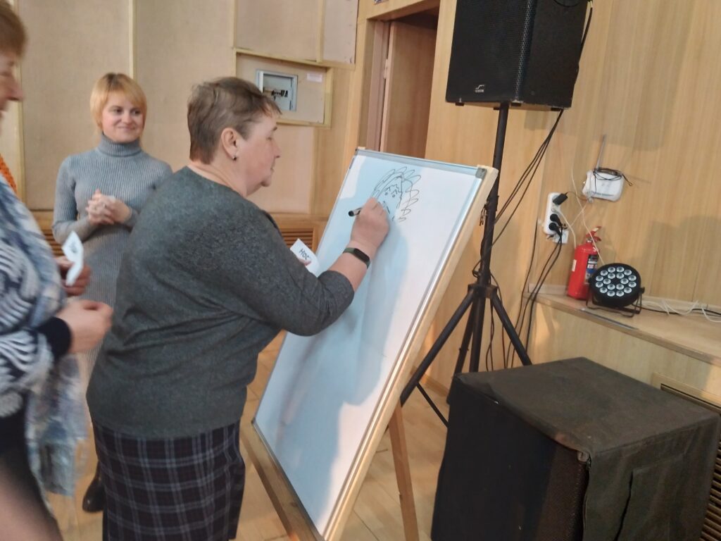 Женщина рисует маркером на наклонённой белой доске; рядом стоят ещё люди и звуковая колонка.