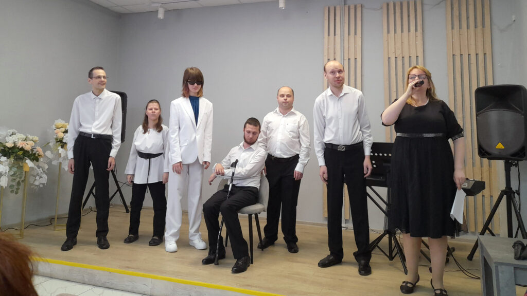 Шесть человек на сцене: пять в белых рубашках/белом костюме и чёрных брюках, один мужчина сидит на стуле с тростью рядом. Справа женщина в чёрном платье и очках держит микрофон и лист бумаги; вокруг колонки и стойки для музыки.