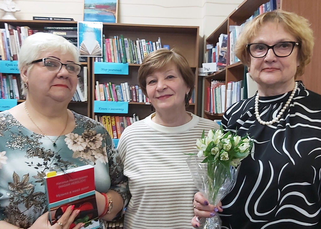 Три женщины в библиотеке на фоне книжных полок: слева в очках с красной книгой, в центре улыбается в светлом полосатом свитере, справа в очках с жемчужным ожерельем и букетом белых цветов