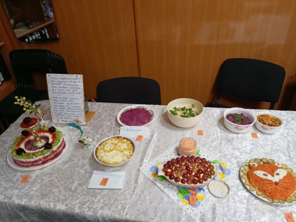 На столе несколько праздничных салатов с табличками.
Есть слоёный салат «тортом», салаты со свёклой, салат с кукурузой и яйцом, блюдо с виноградом и салат, оформленный мордочкой лисы с огурцами по краю.