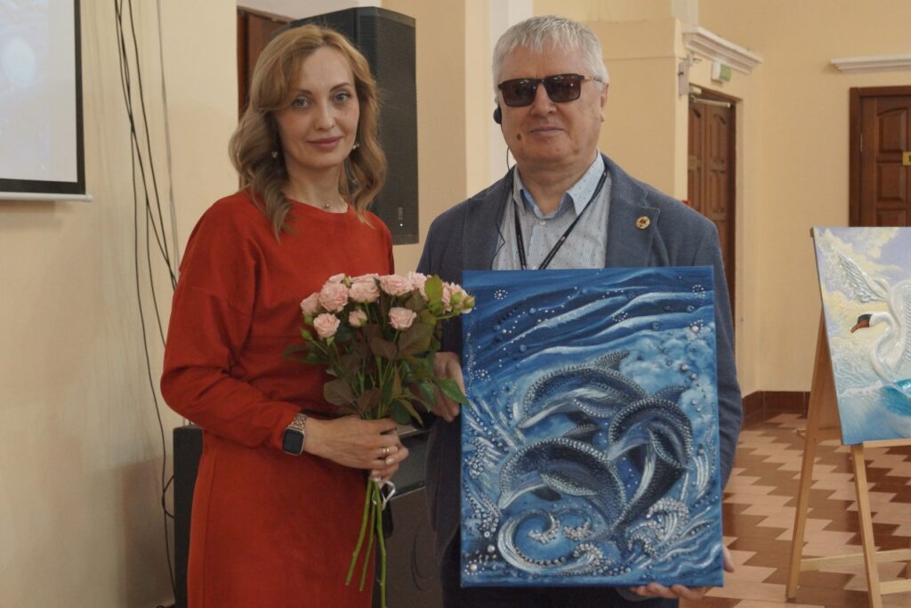 Женщина в красном платье держит букет роз, рядом мужчина в тёмных очках держит сине-голубую картину с дельфинами. В зале; справа на мольберте частично видна картина с лебедем.