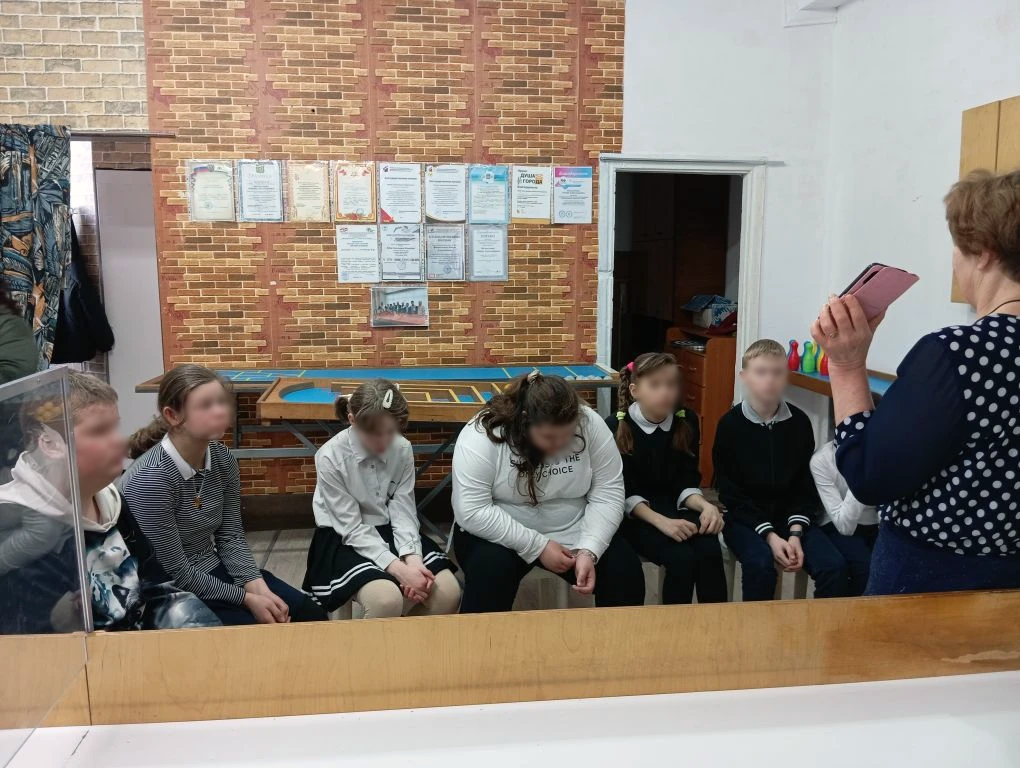 В помещении сидит ряд школьников на стульях, справа стоит женщина с телефоном и обращается к ним; на стене сзади висят грамоты, рядом открытая дверь.