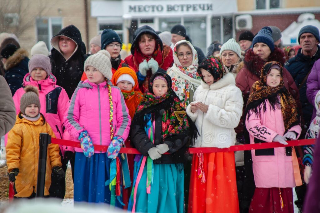 Зимняя уличная толпа за красной лентой; впереди дети в яркой одежде, некоторые в цветных платках и длинных юбках. На фоне здание с вывеской «МЕСТО ВСТРЕЧИ», идёт снег.