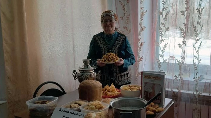 Пожилая женщина в платке и очках стоит за столом с разными блюдами и самоваром, держит тарелку с едой; сзади окно с занавесками.