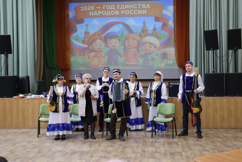 На сцене группа людей в национальных костюмах; в центре мужчина с баяном, рядом женщина с микрофоном, справа мужчина с бас-гитарой. На экране сзади надпись: «2026 — ГОД ЕДИНСТВА НАРОДОВ РОССИИ».