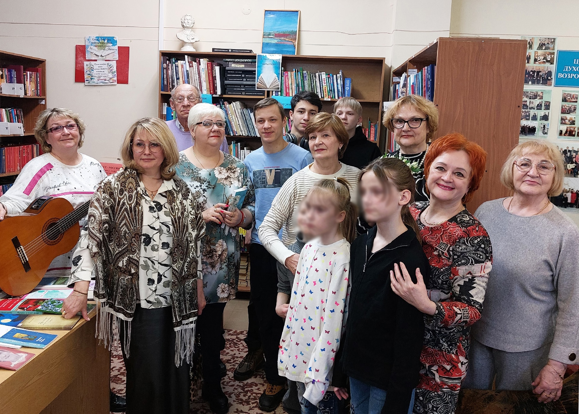 Групповое фото в библиотеке: около 12 человек (в основном женщины, есть молодёжь и двое детей спереди), на фоне книжных стеллажей; слева женщина с гитарой, на столе разложены книги.