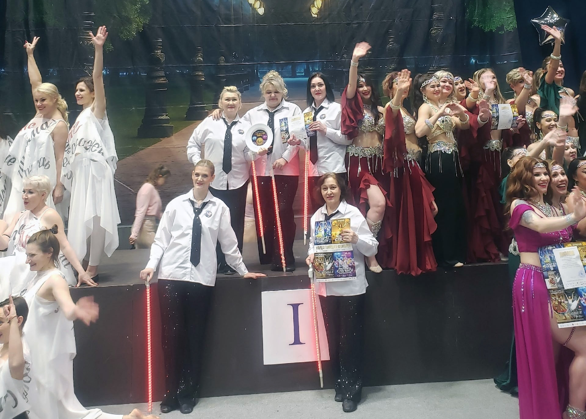 На сцене большая группа женщин после выступления/награждения: в центре несколько в белых рубашках с чёрными галстуками держат дипломы и светящиеся красные палки; справа танцовщицы в бордовых костюмах машут.