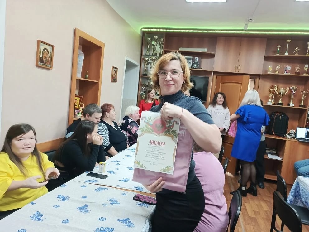 Женщина в очках держит диплом с крупной надписью «ДИПЛОМ» и розовый пакет; вокруг люди за столом, на полках сзади много кубков и наград.