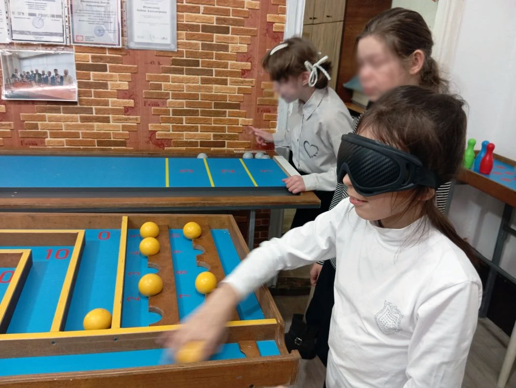 Дети играют в настольную игру с жёлтыми шариками; девочка на переднем плане в повязке на глазах.
