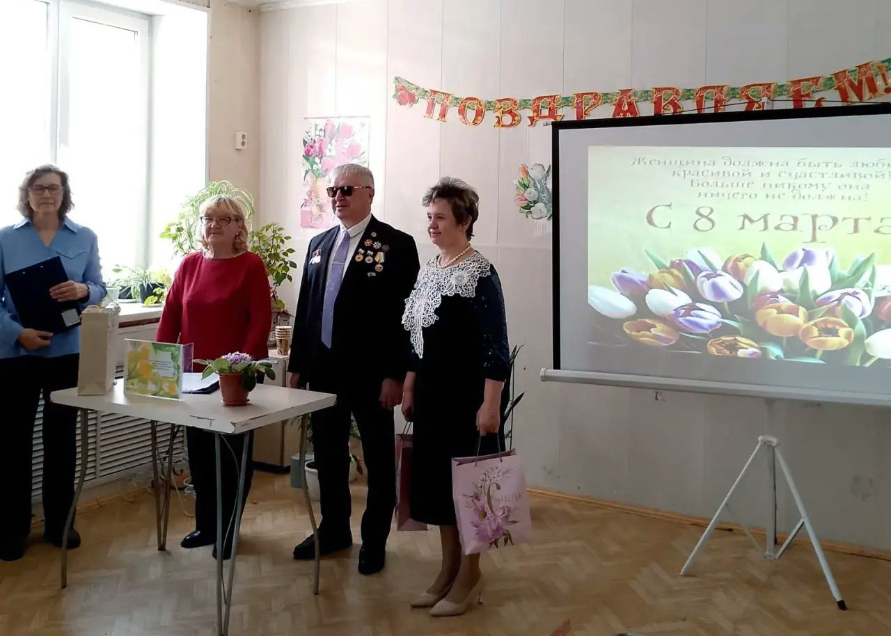 Четыре человека стоят в комнате у экрана проектора с надписью «С 8 марта» и тюльпанами; сверху гирлянда «ПОЗДРАВЛЯЕМ». На столике перед ними открытка и горшок с цветком, у одной женщины в руке подарочный пакет.