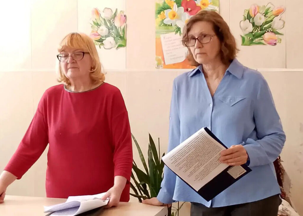 Две женщины в очках стоят у стола: слева в красной кофте, справа в голубой рубашке держит папку с текстом; на стене позади цветочные картинки.