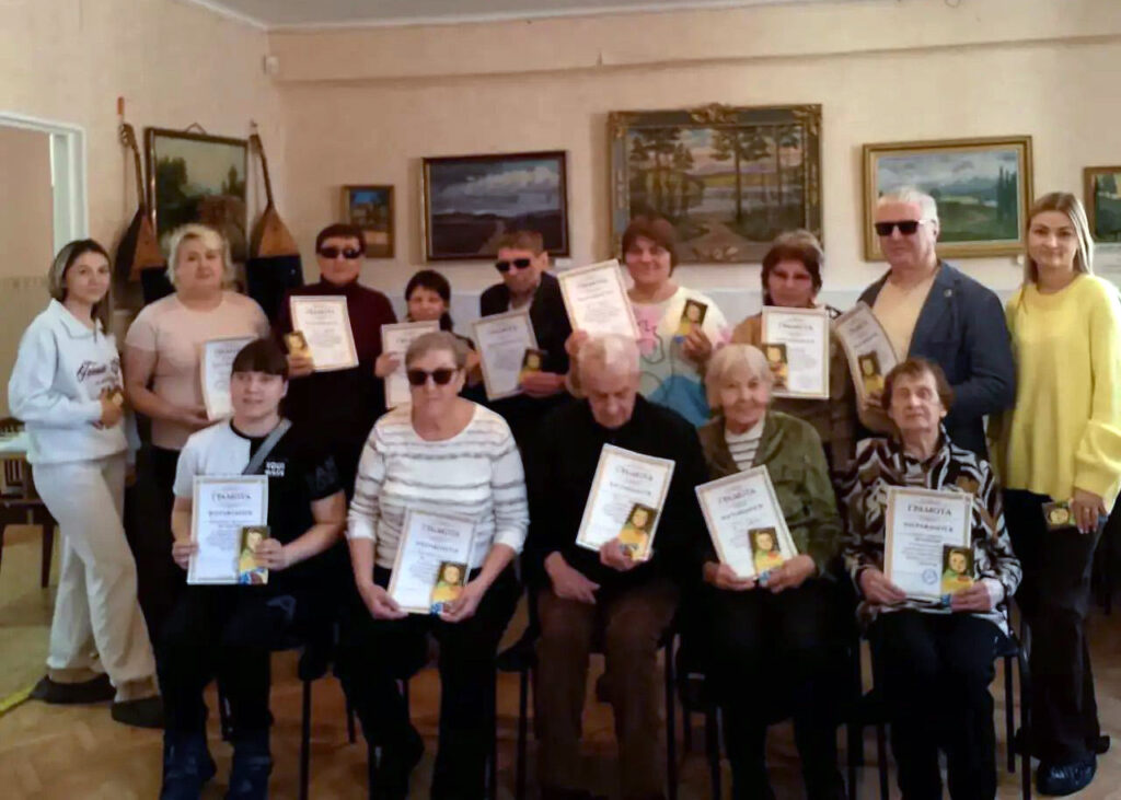 Группа людей в комнате: часть сидит, часть стоит, многие держат дипломы и небольшие книжки; на стене несколько картин, слева висят/стоят балалайки.