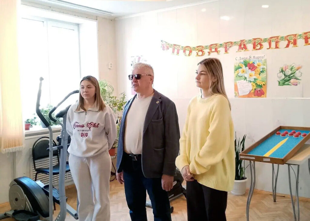 В комнате стоят трое: две девушки и мужчина в тёмных очках. На стене гирлянда «ПОЗДРАВЛЯЕМ» и плакат «С Днём 8 Марта!». Рядом тренажёр и стол с голубой поверхностью и красными шариками.